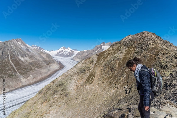 Fototapeta Young Caucasian woman with a backpack on her back looking down at the monumental Aletsch Glacier.This giant stream of ice, which stretches over 23 km