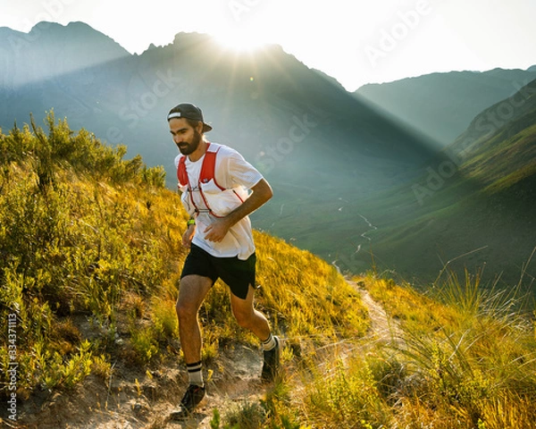 Fototapeta A trail runner at sunrise, running up a mountain trail above a valley in the mountains