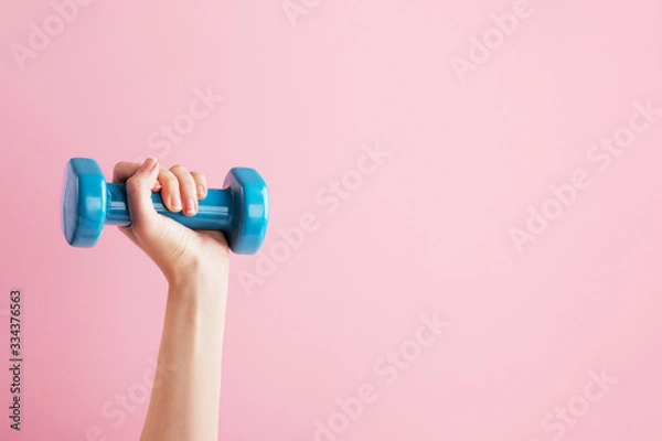 Obraz Womans hand holding blue dumbbell isolated on pink background. Equipment for home workout. Fitness and activity. Sport and healthy lifestyle concept. Copy space in right side