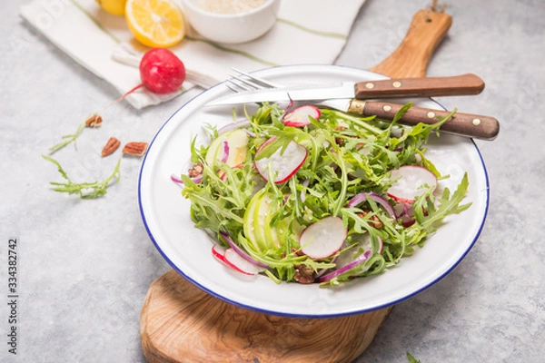 Fototapeta Fresh green arugula leaves on white bowl, rucola rocket salad with apple, radish, pecan nuts, onion on wooden rustic background with place for text. Top view,  healthy food, diet. Nutrition concept