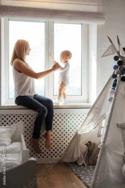 Obraz Portrait of a smiling young cute mother and her son playing near the window in a bright white room. Sunny day outside. True mother love