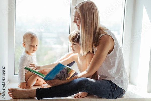 Obraz Portrait of a smiling young cute mother and her sons reading a book near the window in a bright white room. Sunny day outside. 