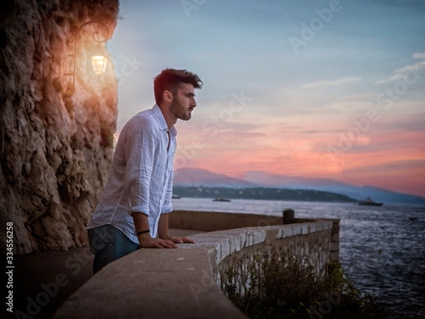 Fototapeta Side view of male in white shirt leaning on border and admiring sea while standing on embankment near rough cliff with lantern against sundown sky