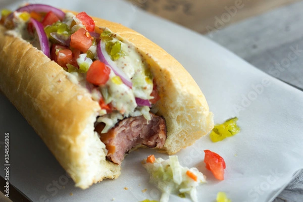 Fototapeta Hot dog with coleslaw, tomatoes, onions and relish. Shown on parchment paper, bite missing. Close view. 