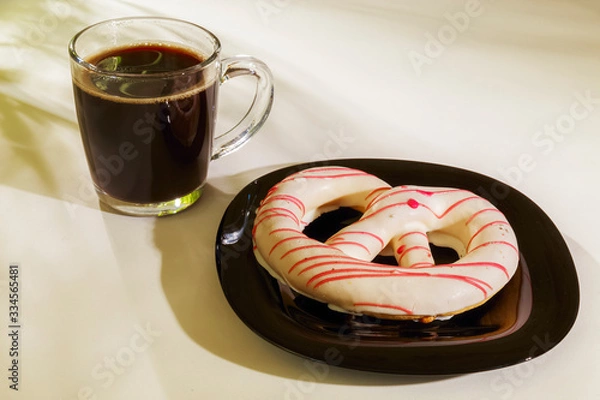 Fototapeta The pretzel cake is on a plate on the table. There is a Cup of coffee next to it. The photo of the Breakfast was taken in the morning light.