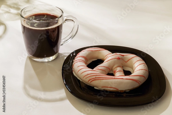 Fototapeta The pretzel cake is on a plate on the table. There is a Cup of coffee next to it. The photo of the Breakfast was taken in the morning light.
