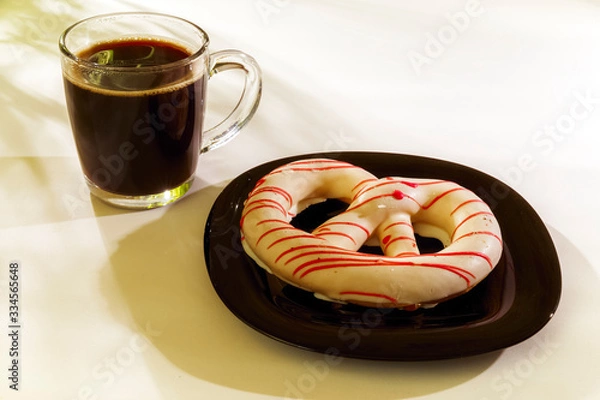 Fototapeta The pretzel cake is on a plate on the table. There is a Cup of coffee next to it. The photo of the Breakfast was taken in the morning light.