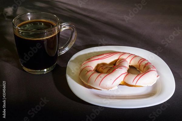 Fototapeta The pretzel cake is on a plate on the table. There is a Cup of coffee next to it. The photo of the Breakfast was taken in the morning light.