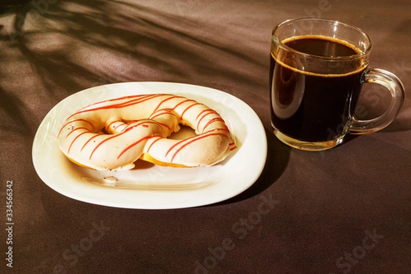Fototapeta The pretzel cake is on a plate on the table. There is a Cup of coffee next to it. The photo of the Breakfast was taken in the morning light.