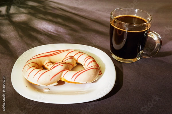 Fototapeta The pretzel cake is on a plate on the table. There is a Cup of coffee next to it. The photo of the Breakfast was taken in the morning light.