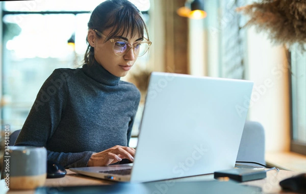 Fototapeta Young business woman in eyeglasses concentrating on screen and typing on laptop while sitting at desk at workplace or cafe. Concept remote work, freelance, using laptop computer.