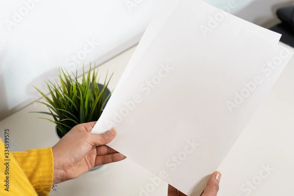 Obraz Close up of a set of two papers hold by a caucasian woman with yellow shirt and bracelet sitting on a desk with a plant