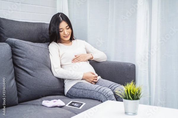 Fototapeta beautiful asian pregnant woman placing hands on baby lump feeling heartbeat smiling joyfully, sitting on sofa relaxing resting from tiredness, in living room with brick texture wall and white curtains