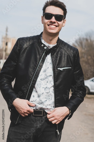 Fototapeta Young happy man in a leather jacket and sunglasses smiling at the camera.. Stylish, cool and brutal guy walks along the street. Portrait of a happy young man. 