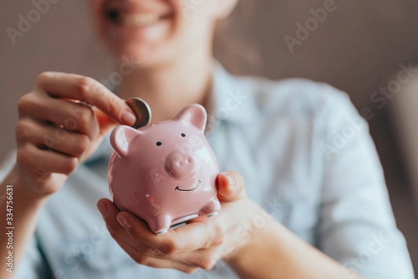 Obraz female hands hold a pink piggy bank and puts a coin there. The concept of saving money or savings, investment
