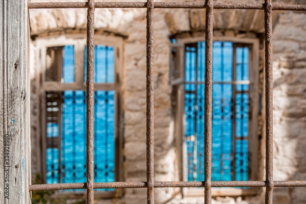 Obraz Rusty wrought iron fence on foreground, two windows with iron fences deeply blurred, turquoise see water surface with ribbles on background