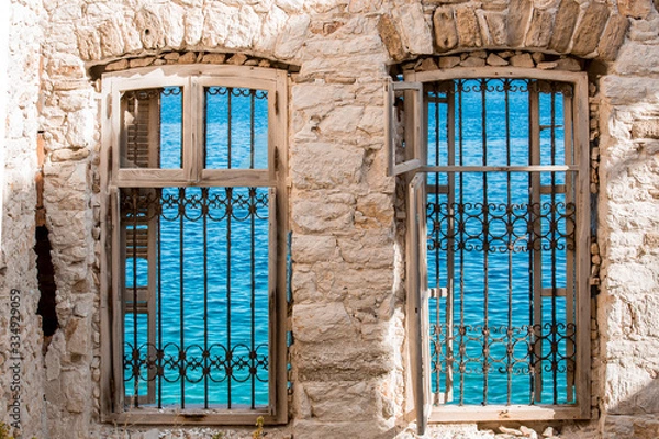 Obraz Two windows with rusty wrought iron fences inside light brown brick house walls, turquoise see water surface with ribbles