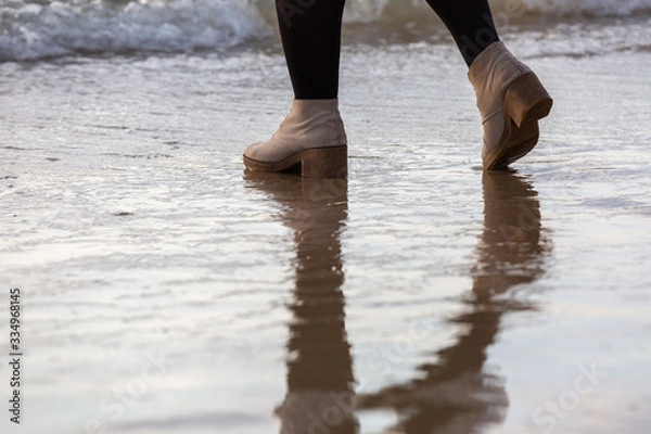 Obraz Girl's legs standing on the beach near the water line in cream-colored winter shoes and black tights, water reflection