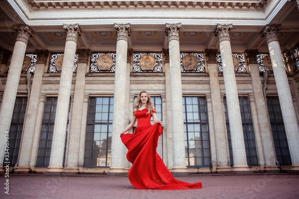 Fototapeta Beautiful blonde girl in a red long dress on a background of architecture, columns 