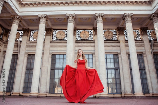 Fototapeta Beautiful blonde girl in a red long dress on a background of architecture, columns 