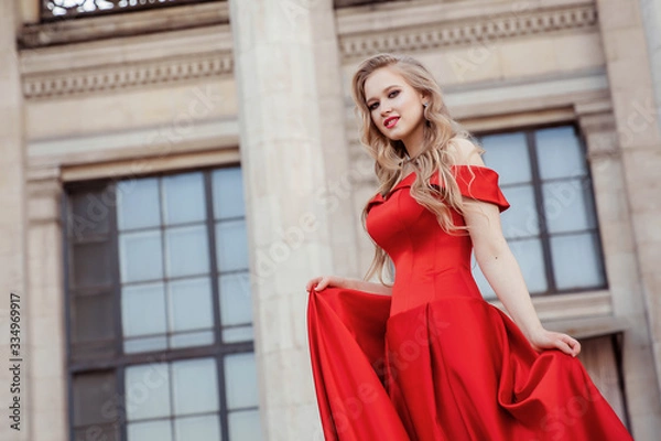 Fototapeta Beautiful blonde girl in a red long dress on a background of architecture, columns 