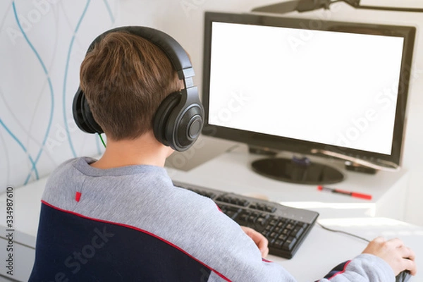 Fototapeta teenager boy looking at computer monitor. computer screen mockup. happy smiling boy in earphones playing computer games. over shoulder view.Distance learning concept.