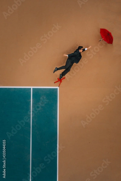 Fototapeta man in a suit reaching for a red umbrella flying away while standing on a red chair on a tennis court