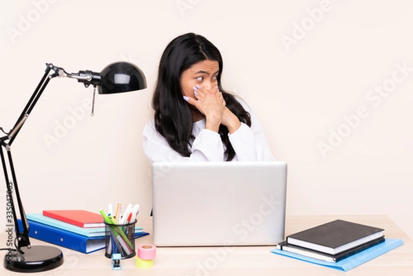Fototapeta Student asian girl in a workplace with a laptop isolated on beige background covering mouth and looking to the side