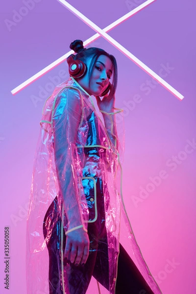 Obraz Beautiful asian woman in a raincoat listens music in headphones around colourful neon