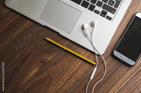 Obraz Espacio de tele trabajo. Vista cenital de ordenador portátil gris, auriculares blancos, lápiz amarillo y negro y teléfono móvil sobre mesa de madera.