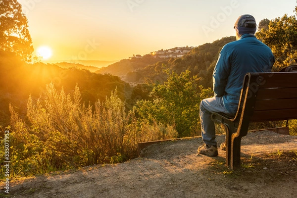 Obraz Person, man, sitting on a bench watching the sunrise over hills and valley with trees and shrubs in the middle and houses on a hill in the background