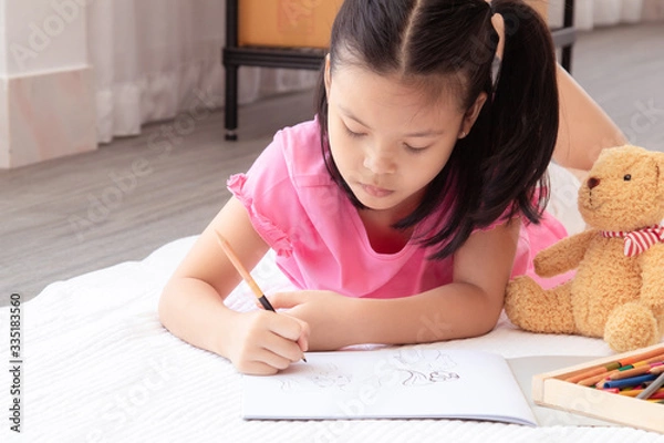 Fototapeta Asian home school elementary adorable kids lie down on floor, drawing homework with color pencils, happy cheerful girl spent time concentrate  drawing quarantine period before back to school