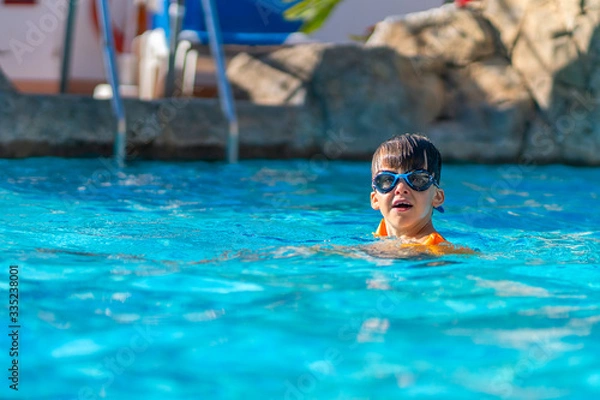 Fototapeta A happy boy in swimming goggles and arm ruffles in an outdoor pool. The child learns to swim. Family holidays at a tropical resort.
