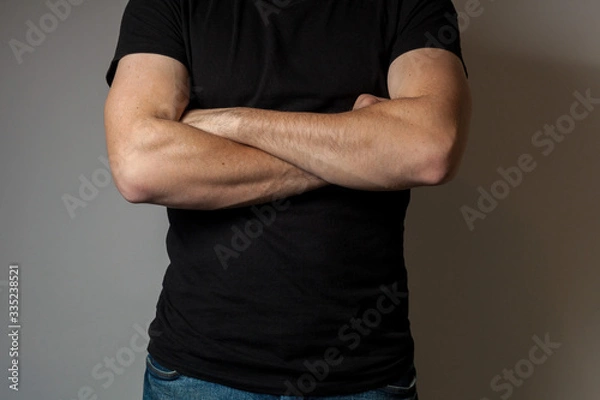 Fototapeta A man in a black T-shirt crossed his arms in front of his chest against a gray wall background
