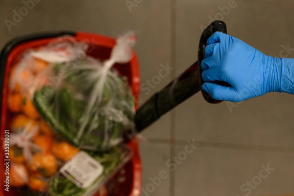 Fototapeta A basket of products in a store. A female hand holds it in a blue protective glove.The concept of protection and disinfection in times of viruses,epidemics, pandemics. Stock of products for quarantine