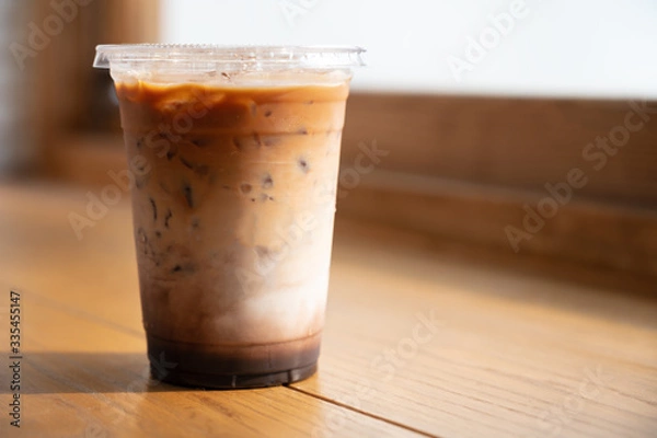 Fototapeta Iced caramel macchiato in plastic take away cup on wooden table near the window in the morning with copy space.