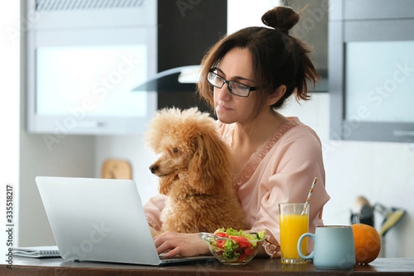 Fototapeta The young woman is working remotely. Young woman with her dog working using a laptop at home. Concept of the workplace at home, working remotely.