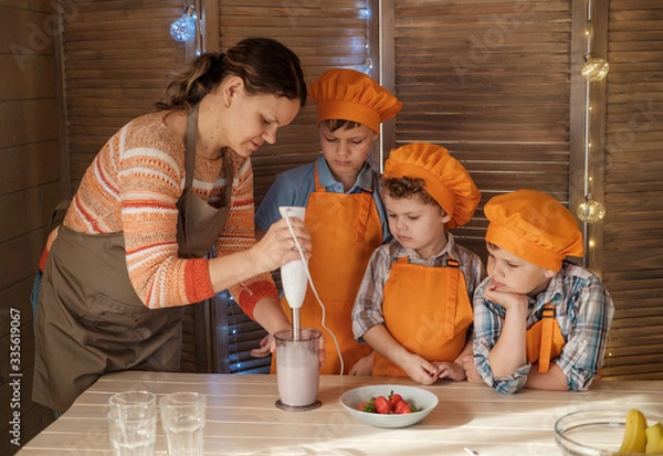 Fototapeta a mother and three sons prepare a milkshake with strawberries and a banana in the kitchen. boys dressed as cooks prepare dessert with their mother