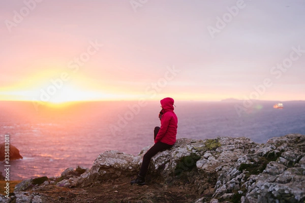 Fototapeta Woman sitting on the rocks watching the sunset in Martins Haven with St. Brides Bay in the background in Pembrokeshire, Wales, UK.