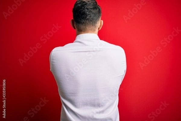 Fototapeta Young business man with blue eyes wearing elegant shirt standing over red isolated background standing backwards looking away with crossed arms