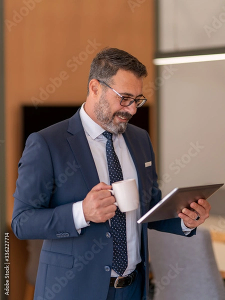 Obraz An office worker who actively uses social media, checking incoming data from his tablet during his standing coffee break