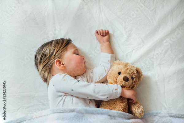 Fototapeta Toddler baby sleeping on white sheets hugging soft brown teddy bear. Children's nap time or bedtime. Child sleep deprivation. Child sleep routine and schedule.  Top view. 