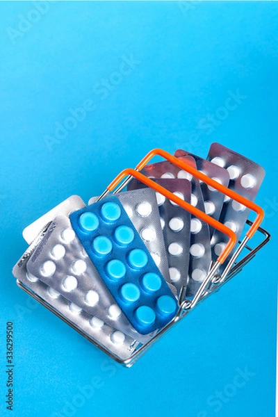 Fototapeta A small shopping basket filled with blisters of tablets on a blue background. Concept for the theme of the sale / purchase of drugs.