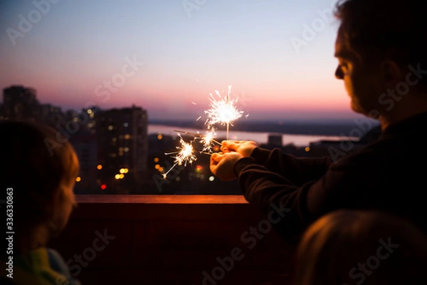 Fototapeta Family dad and two kids with Sparklers At sunset on the balcony of the home. Corona Virus. standing in home. chelter in place. end of a quarantine. home holiday
