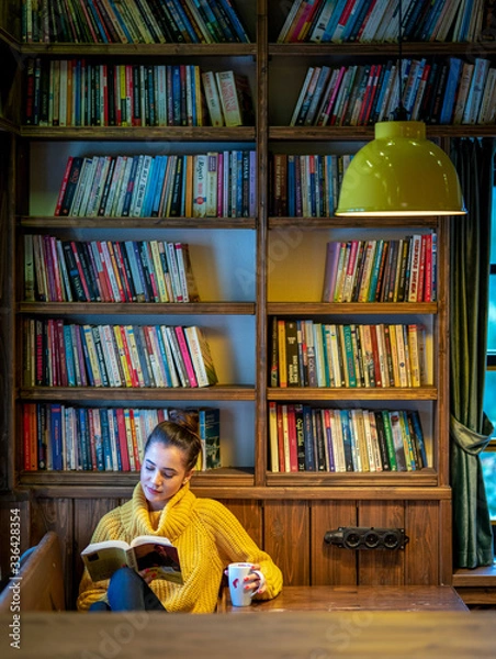 Obraz On the couch in front of the window, a woman caught up in reading. The woman at the table drinking coffee and reading a book