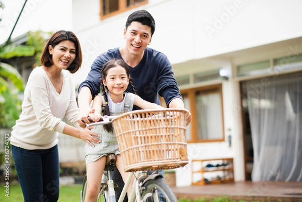 Obraz Young Asian family love, father and mother teach and training daughter to ride a bike on grass field at outdoor of home. girl train to ride a bike by her parent which smiling and felling happy