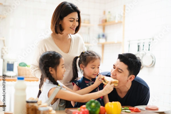 Fototapeta Young Asian love family are preparing cooking the breakfast, sandwich vegetable on table in the kitchen which Excited smiling and felling happy. daughter feed meal to father