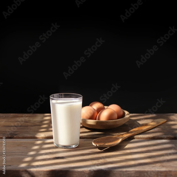Fototapeta brown eggs lie on a wooden bowl, an old spoon and a glass of white milk in the rays of light on the table, retro concept