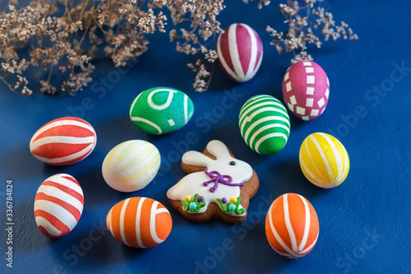 Fototapeta The attributes of the chrestian Easter holiday are colored eggs and gingerbread in the form of a rabbit on a dark blue table