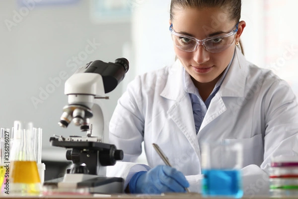 Fototapeta Portrait of serious researcher looking at camera with calmness. Smiling laboratory assistant writing research data in documents. Chemical experiments concept